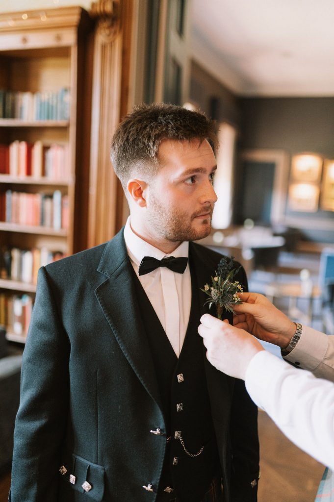 Man in a black suit and bow tie has a boutonniere pinned to his lapel in a wood-paneled room, likely preparing for a formal event.