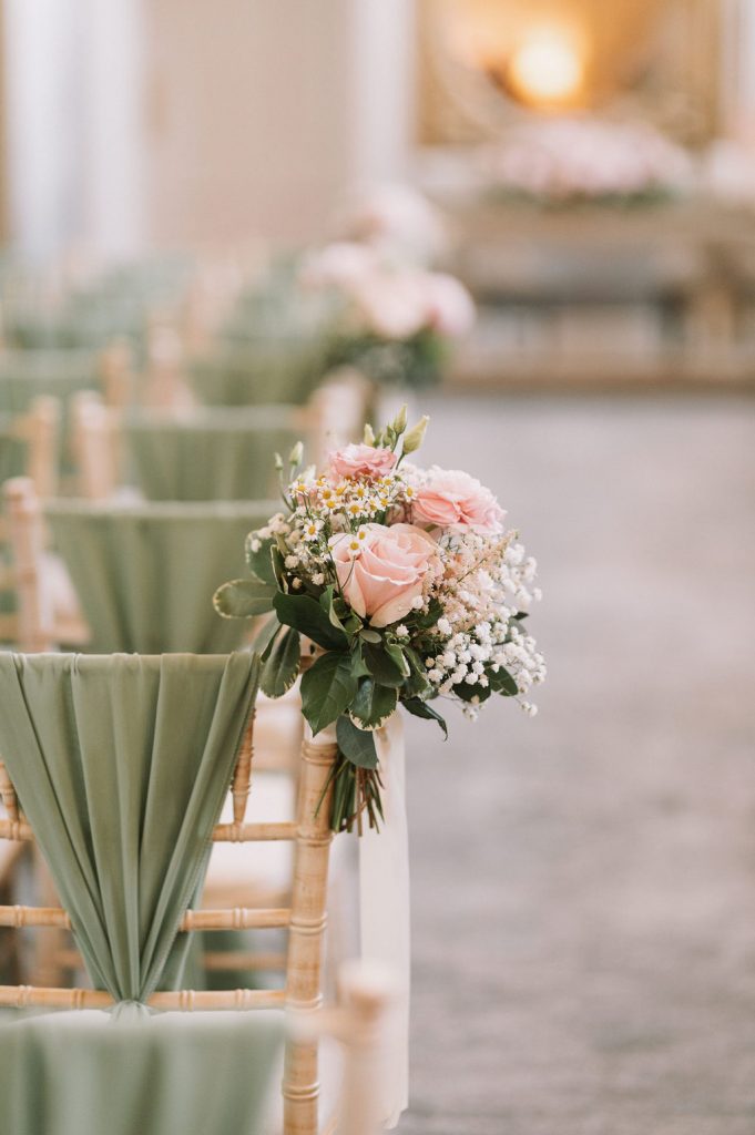 Decorated ceremony chairs with sage-green fabric and pastel pink bouquet of roses and baby's breath tied to the back.