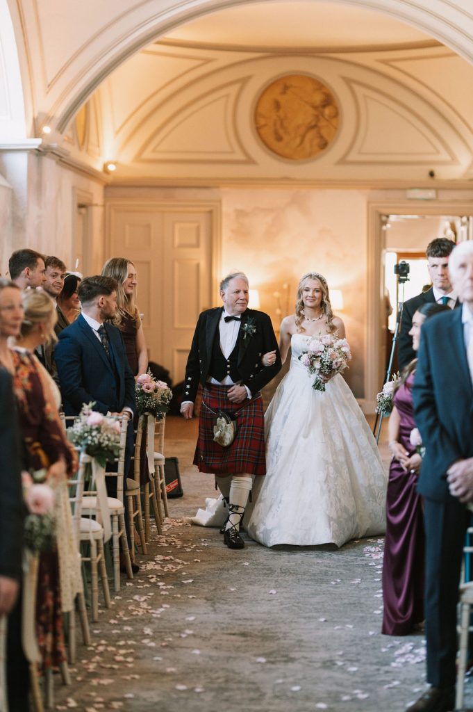 Bride in a white gown carries a bouquet as she is escorted down an aisle by a man in a formal tartan kilt; guests sit on both sides in a warmly lit hall.
