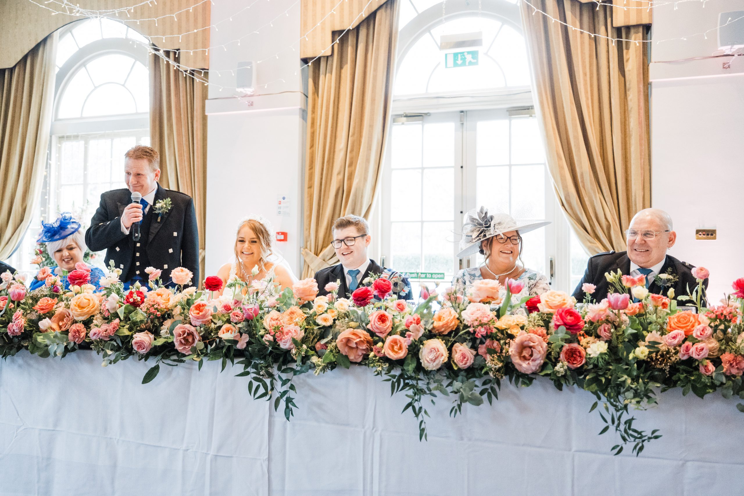 balbirnie-house-wedding-fife-fraoch-floral-design - Fraoch Floral Design Wedding reception head table adorned with a dense bed of pink, peach, and coral flowers; guests smile in the background, one man speaks into a microphone.