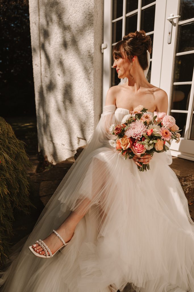 Bride in off‑the‑shoulder white wedding gown holding a pink and peach bouquet, standing by a glass door.