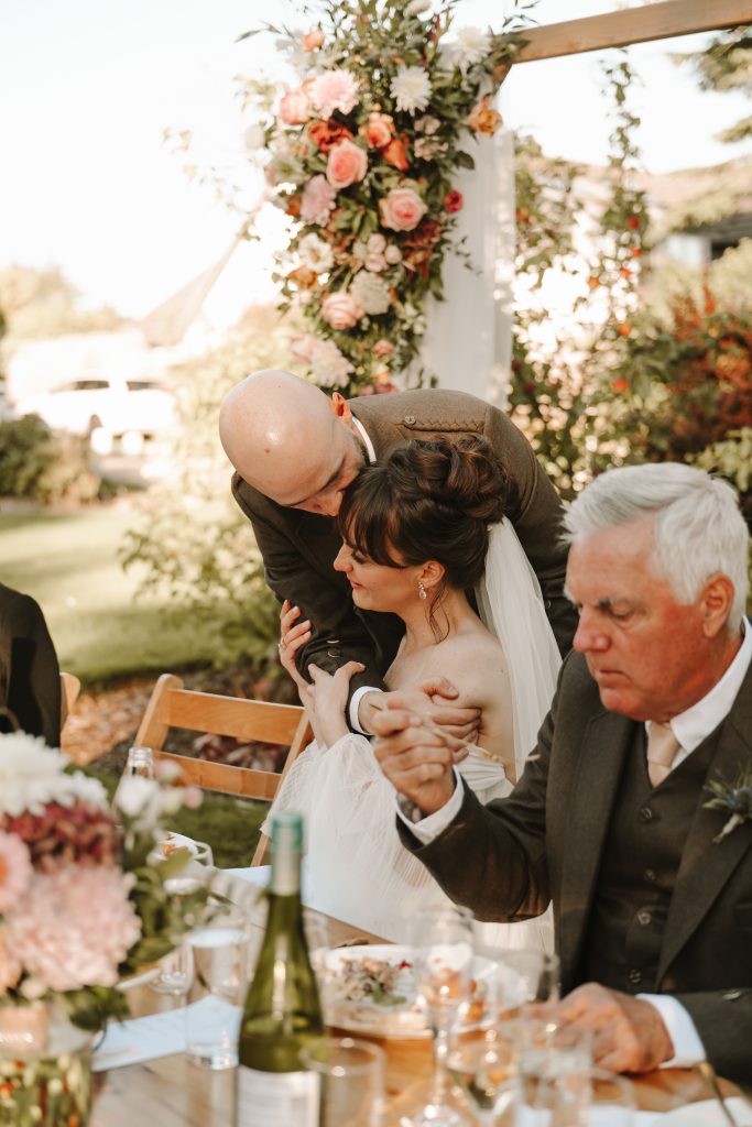 Bride seated at a wedding table while a man hugs her from behind during an outdoor ceremony, with a floral arch in the background.