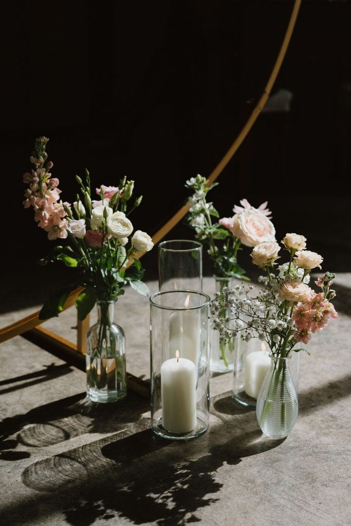 Floral centerpiece on a table with pink and white blooms in glass vases and tall candles, cast in dramatic shadows on a dark background.