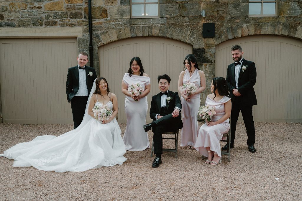 Bride in a white gown with train, groom in a tux, and bridesmaids in blush dresses holding bouquets, posed before a stone building with arches.