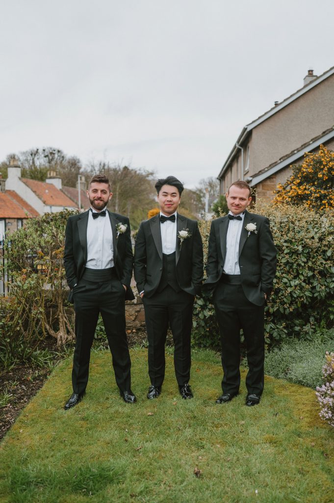 Three men in black tuxedos with bow ties posing outdoors in a garden, boutonnières on their lapels.
