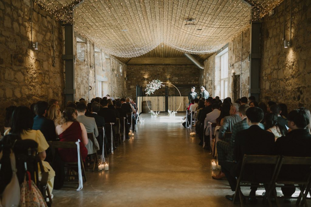 Wedding ceremony in a rustic stone hall with string lights and a circular floral arch at the front, guests seated on both sides.