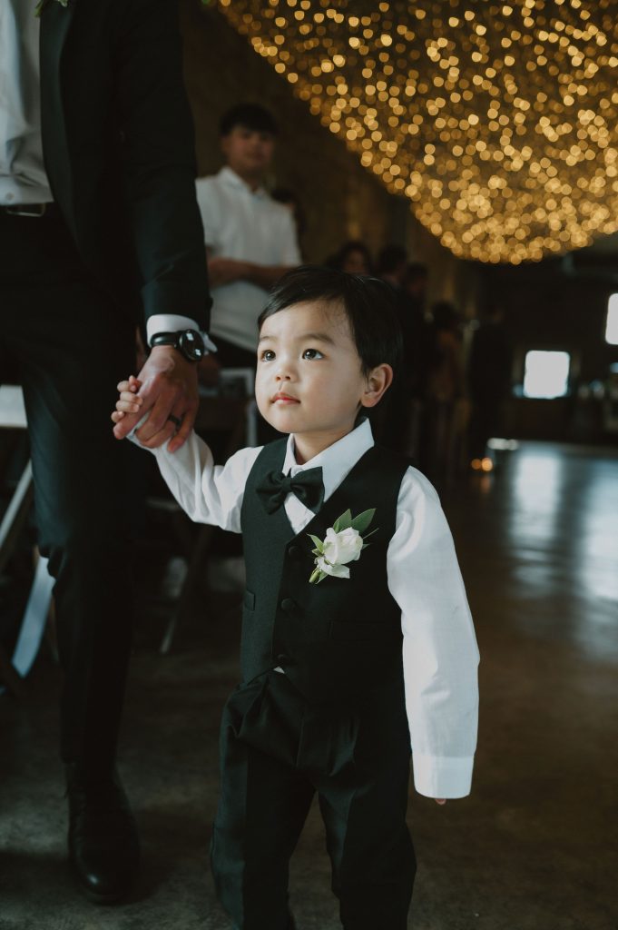 Young boy in a black vest and bow tie, holding an adult's hand at a wedding with blurred string lights behind.