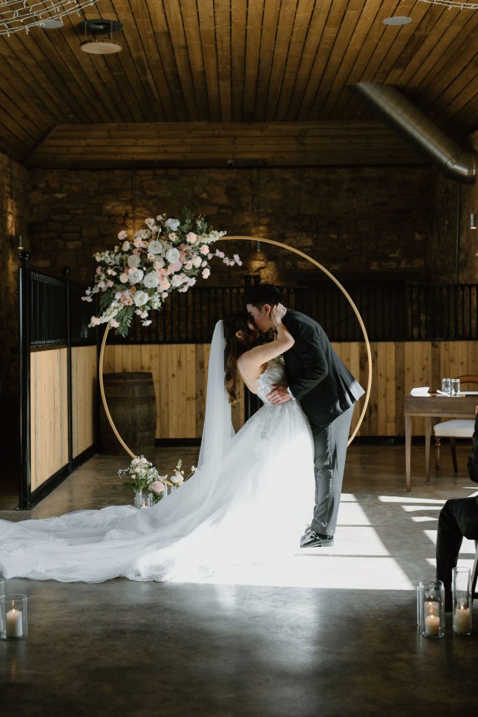 A bride and groom kiss beneath a circular floral arch in a rustic indoor venue, candles on the floor nearby and a long white wedding gown trail.