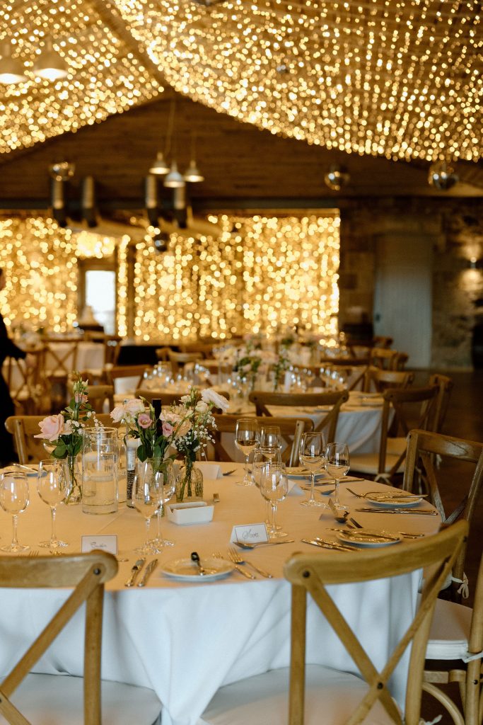 Elegant wedding reception setup with white tablecloths, floral centerpieces, and warm string lights overhead.