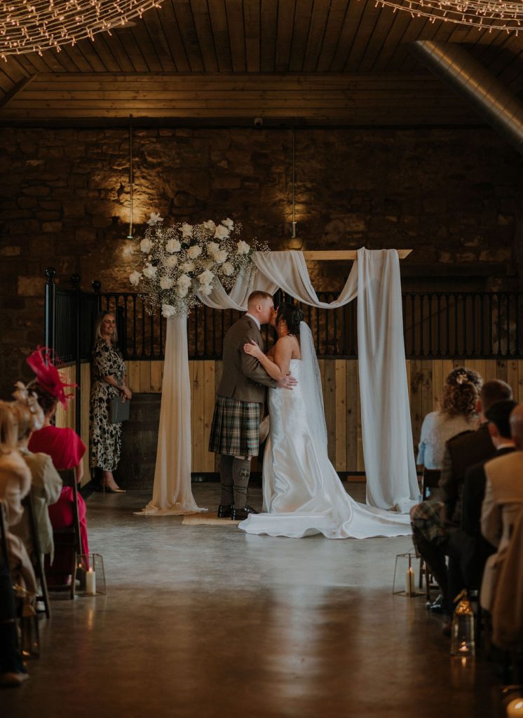 Bride and groom kiss beneath a draped floral arch during a rustic indoor wedding, with guests seated on either side and string lights above.