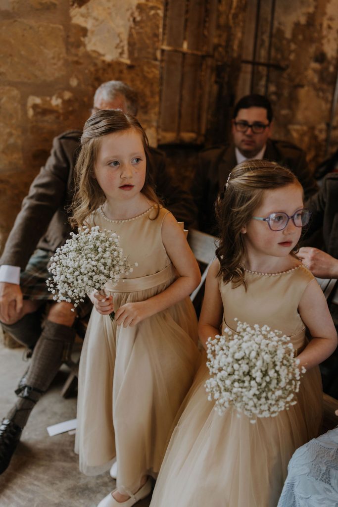 Two young flower girls in champagne dresses stand with bouquets of baby’s breath at a rustic ceremony venue, watching attentively.