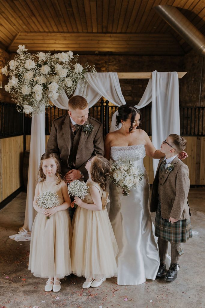 Bride and groom pose with two flower girls in cream dresses and a boy in a tartan kilt under a white floral arch in a rustic venue.