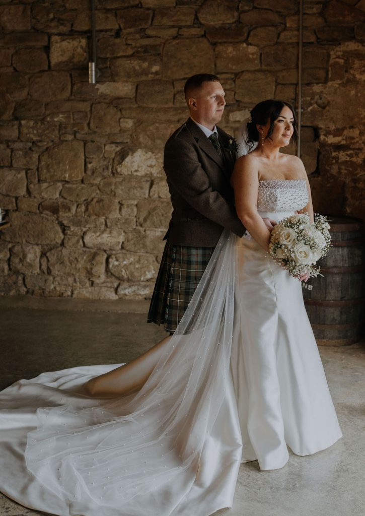 Bride in a white strapless gown with a long beaded veil and bouquet of white roses stands beside a groom in a dark jacket and kilt against a stone wall.
