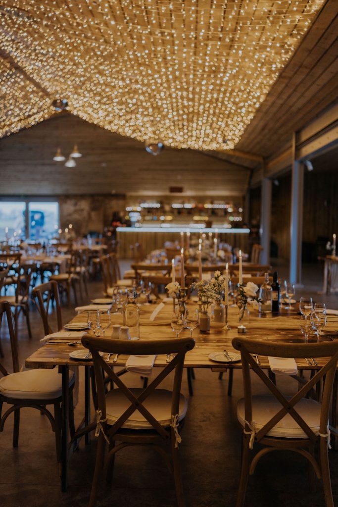 Elegant rustic dining hall with long wooden tables set for a formal dinner, candles, flowers, and twinkling string lights overhead.