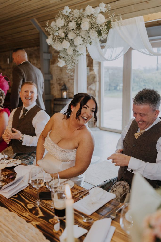 Bride in a beaded strapless gown laughing at a rustic wedding reception, seated with two men in vests at a candlelit wooden table.