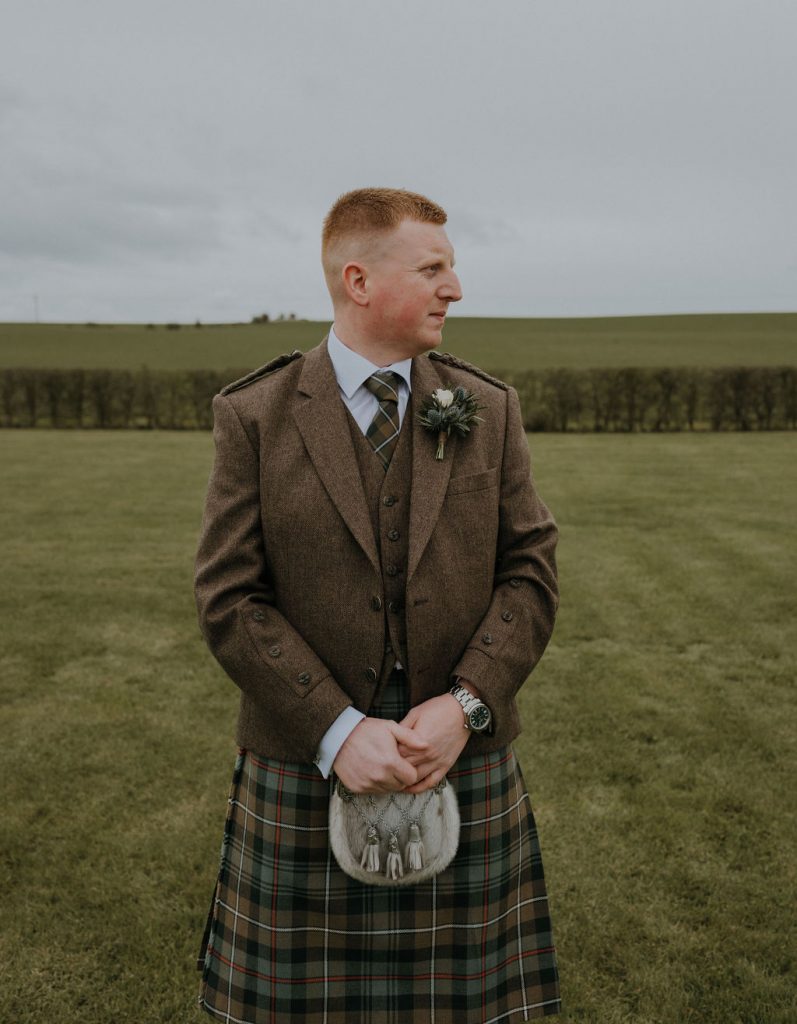 Man in a brown tweed jacket and plaid kilt stands in a grassy field, looking to the right, with a boutonniere on his lapel.