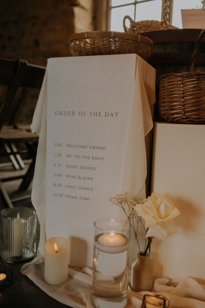 Wedding program sign titled 'Order of the Day' with a schedule (Welcome Drinks, We Tie the Knot, More Drinks, Wine & Dine, First Dance, Home Time) placed on a draped white cloth, surrounded by candles and flowers in a cozy setting.