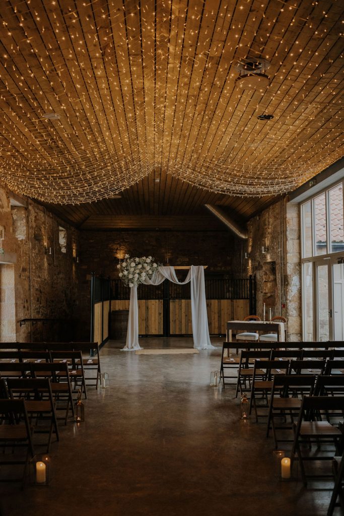 Wedding ceremony space in a rustic hall with twinkling string lights overhead, a white fabric arch, and rows of wooden chairs by candlelit aisles.