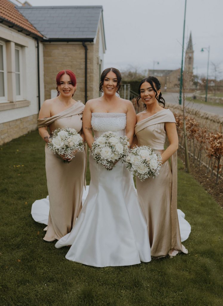 Bride in a white strapless gown stands between two bridesmaids in champagne dresses, all holding white bouquets outdoors.