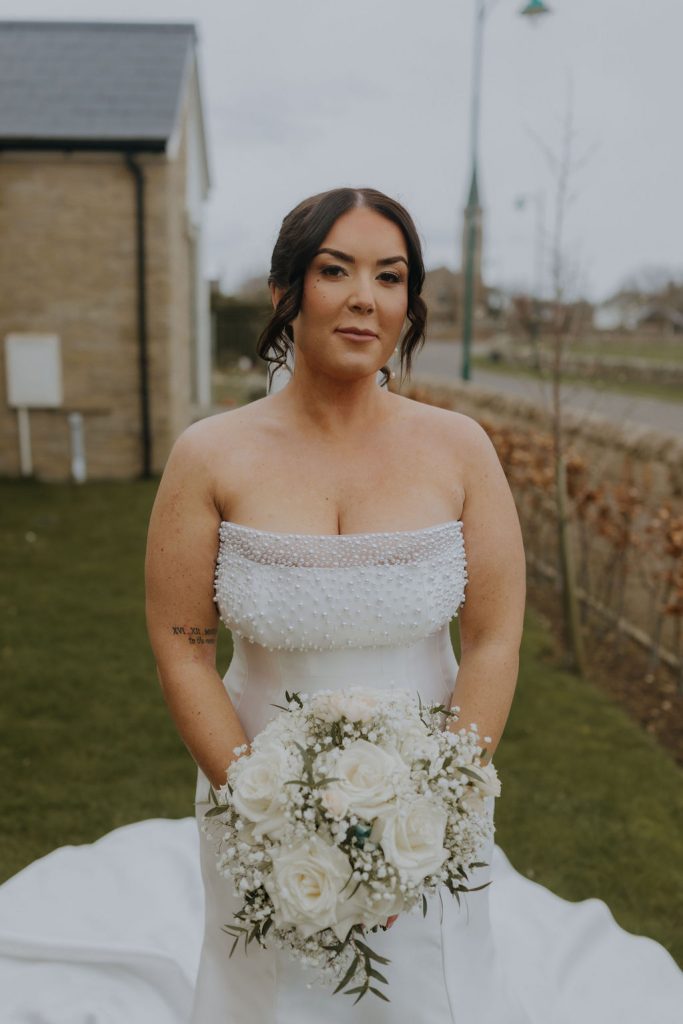 Bride in a strapless white wedding gown with pearl embellishments, holding a white bouquet outdoors.