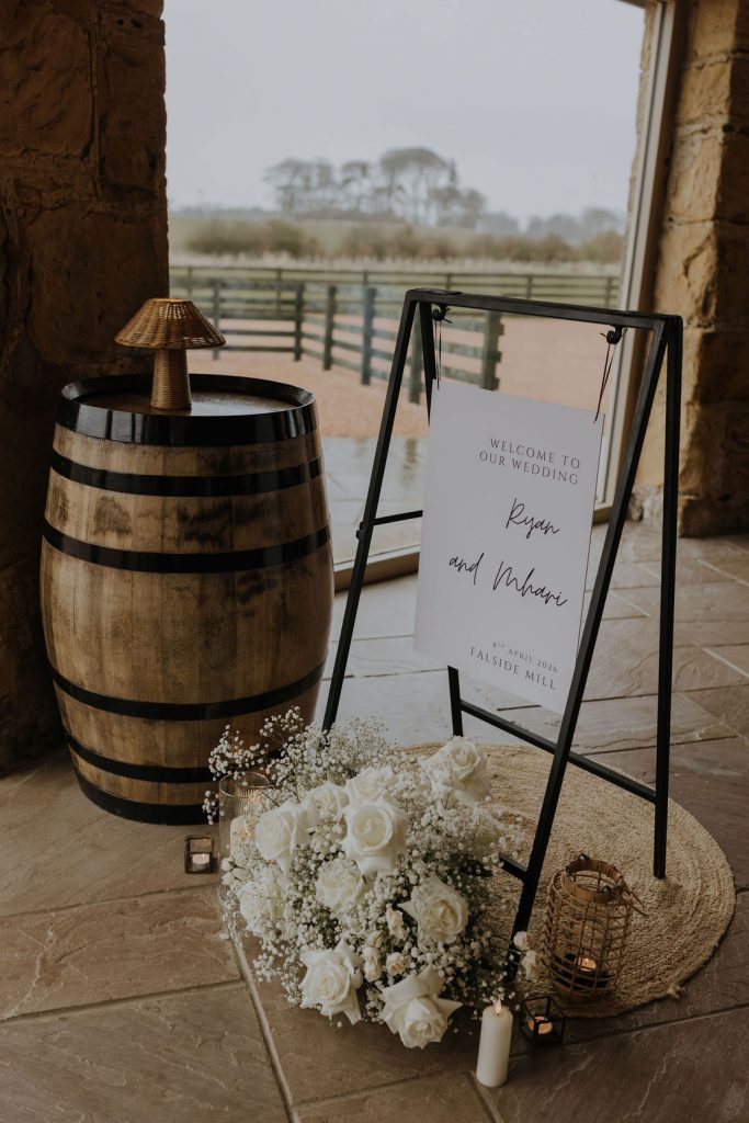 Wedding welcome sign on a black metal stand beside a wooden barrel, with a white floral bouquet and candles on the floor, in a rustic venue.