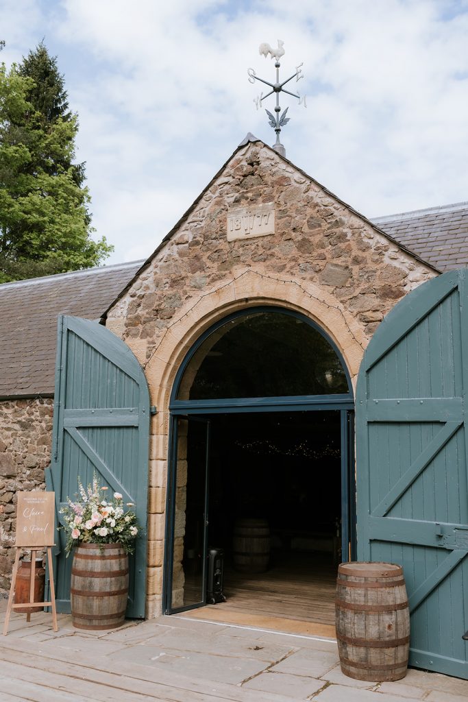 Paul Claire - Fraoch Floral Design Rustic wedding venue exterior with a stone archway and teal double doors open to a warm interior, floral arrangements in a barrel outside and a wooden sign nearby.