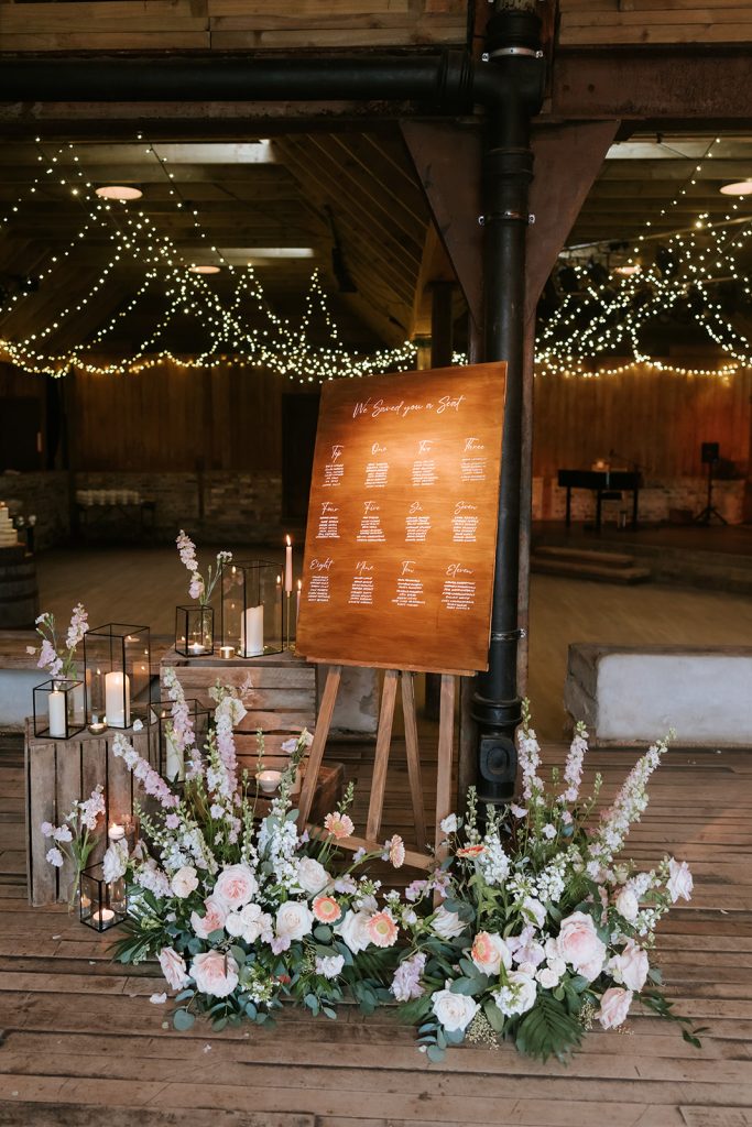 Paul Claire - Fraoch Floral Design Wooden wedding seating chart on an easel surrounded by pink and white flowers and lanterns in a rustic venue.