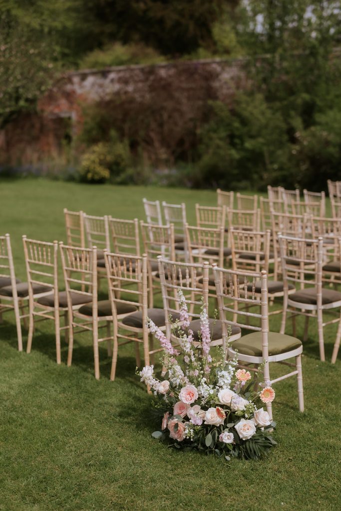Paul Claire - Fraoch Floral Design Rows of beige chiavari chairs arranged on a lawn for an outdoor wedding, with a pastel floral centerpiece at the front