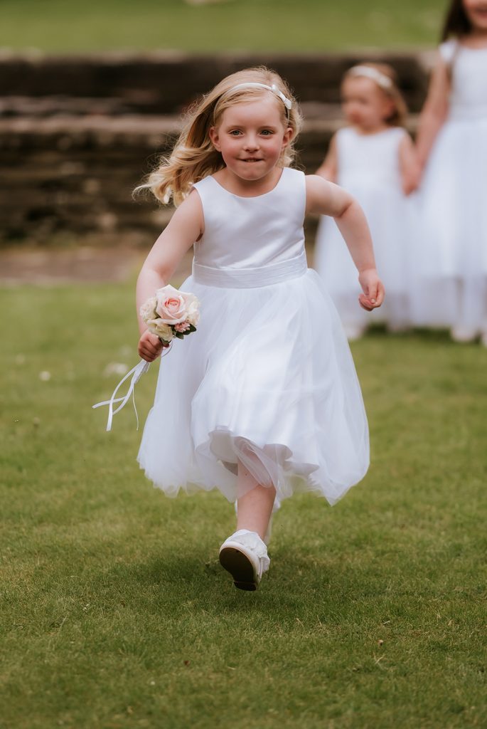 Paul Claire - Fraoch Floral Design Flower girl in a white dress runs on a grass lawn, holding a pink bouquet.