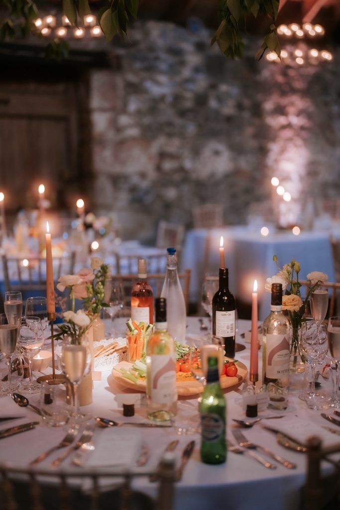 Paul Claire - Fraoch Floral Design Table set for a celebration with candles, wine bottles, flowers, and a veggie platter against a stone wall.