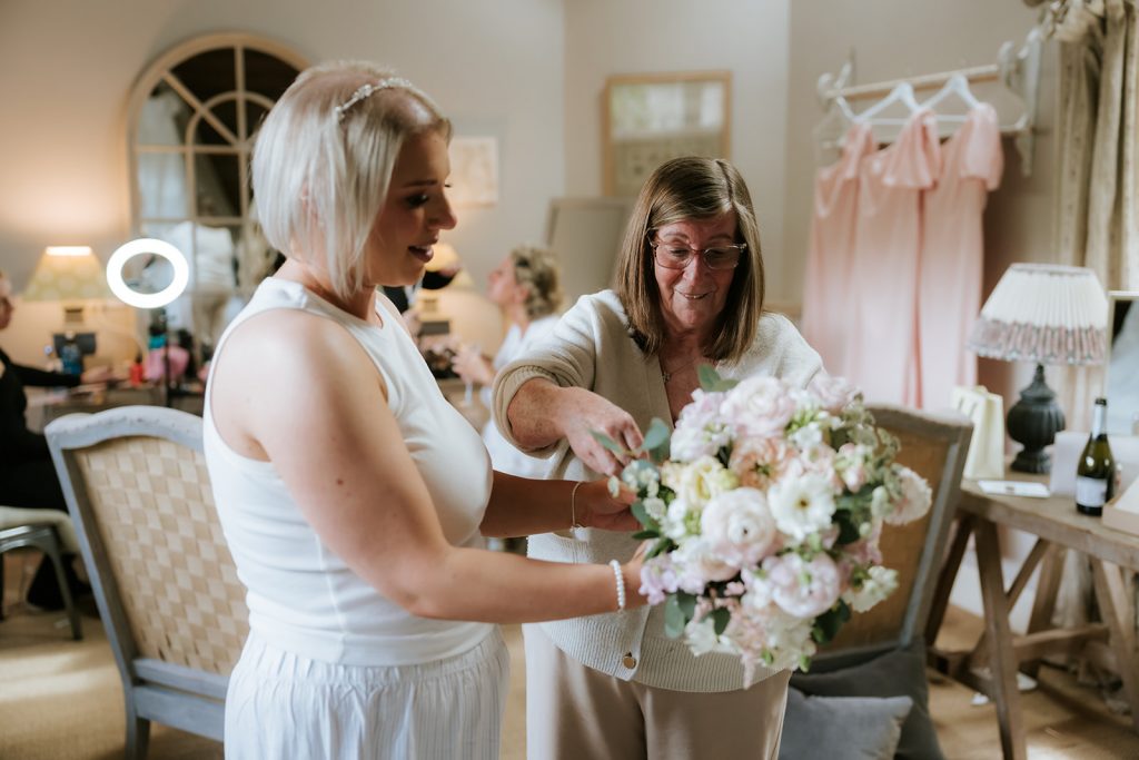 Paul Claire - Fraoch Floral Design Bride-to-be in a white dress hands a bouquet to an older woman in a bridal shop, surrounded by pink dresses and styling setup.