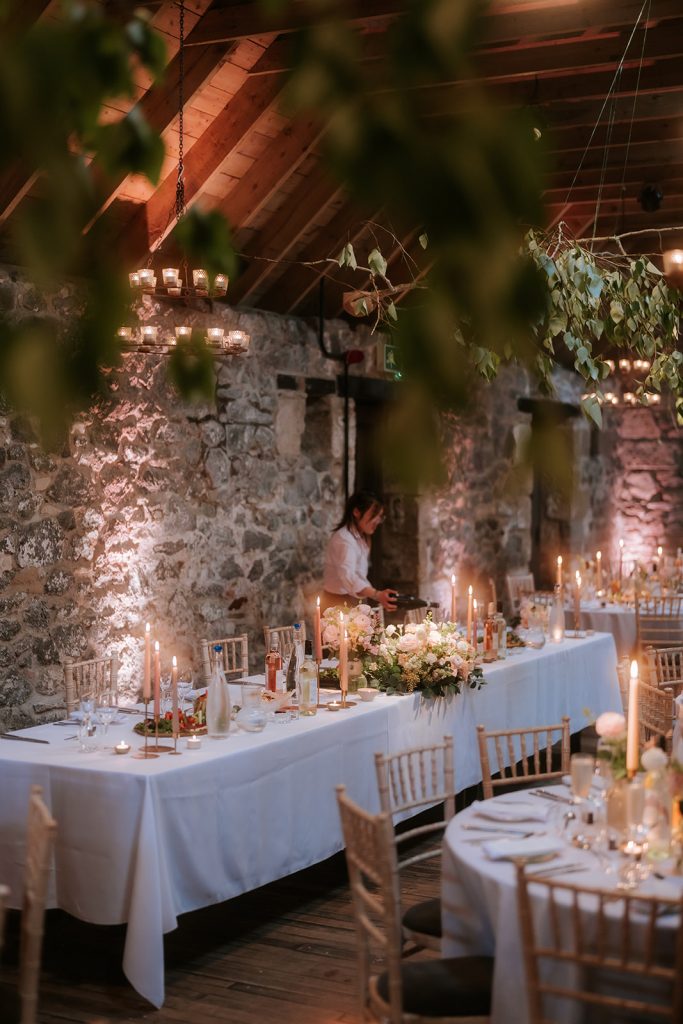 Paul Claire - Fraoch Floral Design Rustic wedding reception in a stone-walled room, with long white-clothed tables, candles, and floral centerpieces.