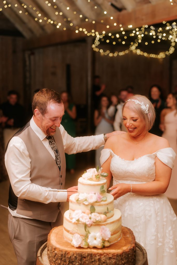 Paul Claire - Fraoch Floral Design Bride and groom smile as they cut a layered wedding cake decorated with flowers. Rustic venue with string lights in the background.