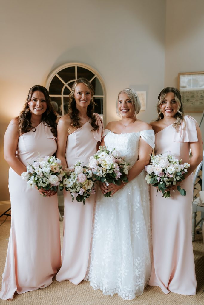 Paul Claire - Fraoch Floral Design Bride in a white lace gown flanked by three bridesmaids in blush pink dresses, all holding bouquets of white and pink flowers inside a warmly lit room.