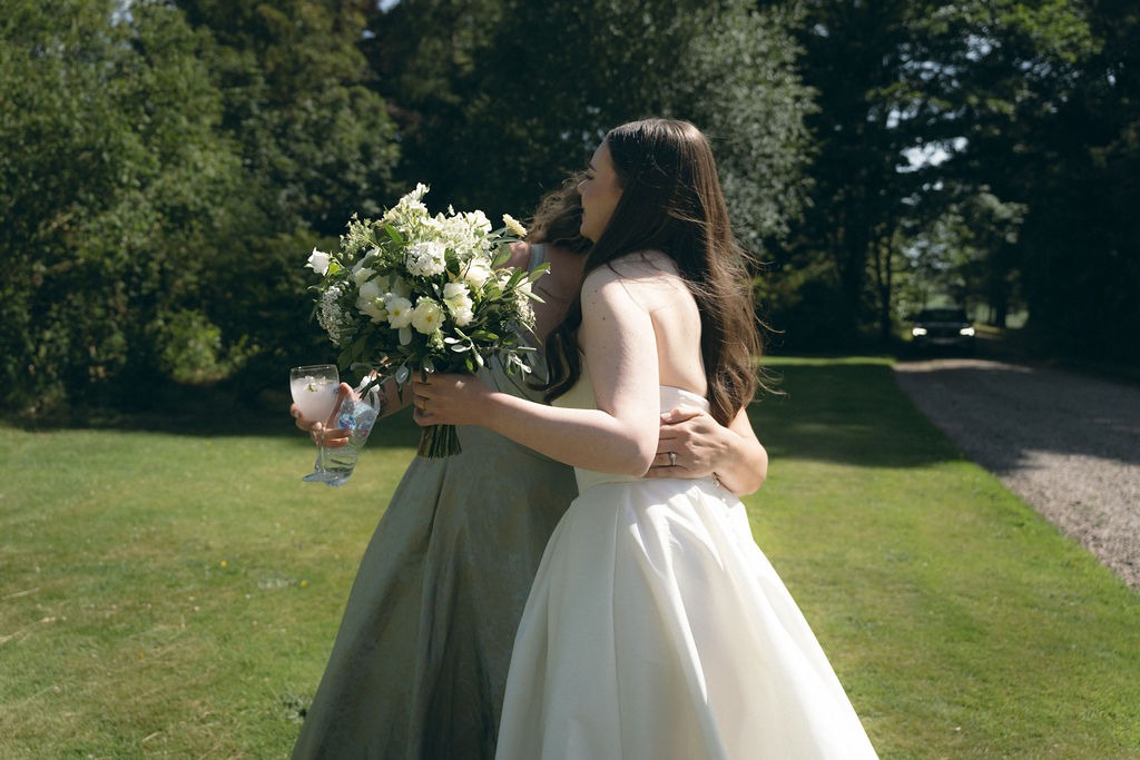 kinblethmont-house-wedding-angus-fraoch-floral-design 12 - Fraoch Floral Design Bride in a white wedding dress hugs a bridesmaid outdoors, holding a bouquet of white flowers (and a glass).