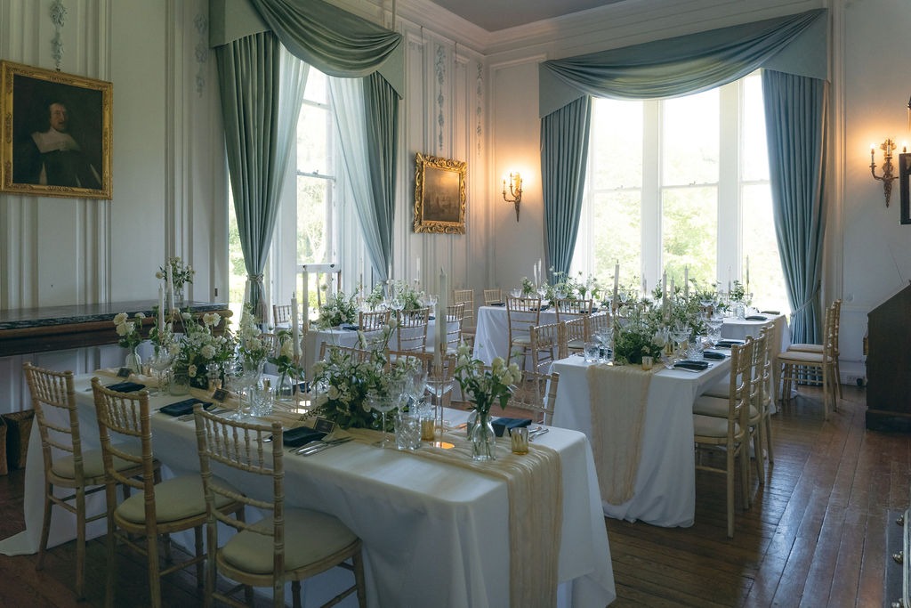 kinblethmont-house-wedding-angus-fraoch-floral-design 14 - Fraoch Floral Design Elegant dining room set for a formal event with white tablecloths, cream runners, gold chairs, and floral centerpieces by tall windows.