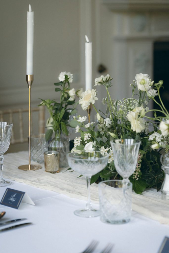 kinblethmont-house-wedding-angus-fraoch-floral-design 17 - Fraoch Floral Design Elegant dining table with white flowers, tall tapered candles in brass holders, and crystal glassware on a white tablecloth.