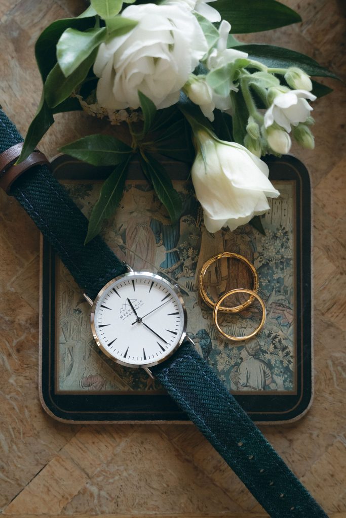 kinblethmont-house-wedding-angus-fraoch-floral-design 3 - Fraoch Floral Design White bouquet with green leaves, gold rings, and a silver-faced watch arranged on a vintage tin box with a dark strap nearby.
