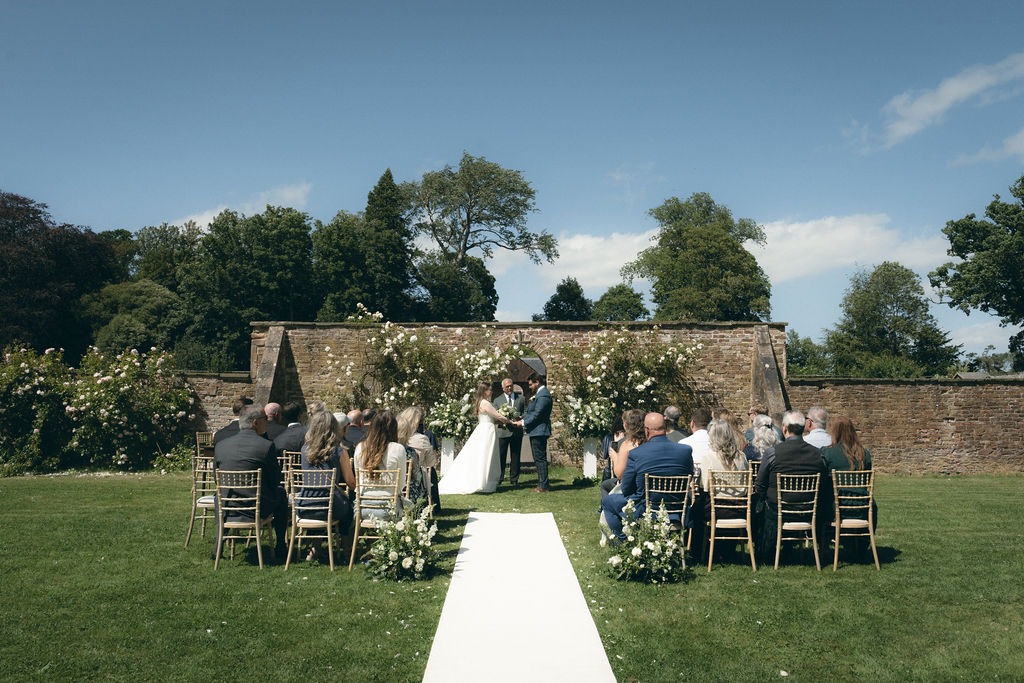 kinblethmont-house-wedding-angus-fraoch-floral-design 7 - Fraoch Floral Design Outdoor wedding ceremony with couple exchanging vows under floral arch on a white aisle runner, guests seated on wooden chairs in a garden setting.