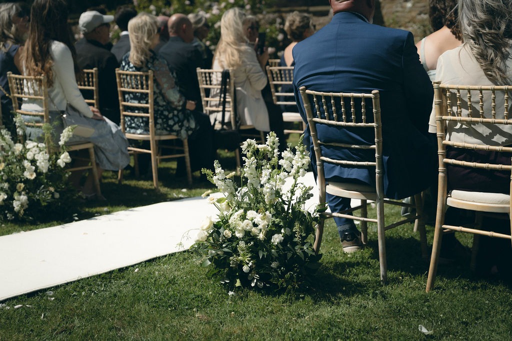 kinblethmont-house-wedding-angus-fraoch-floral-design 8 - Fraoch Floral Design Outdoor wedding ceremony with white floral arrangements along a grass aisle and guests seated in gold Chiavari chairs outside.