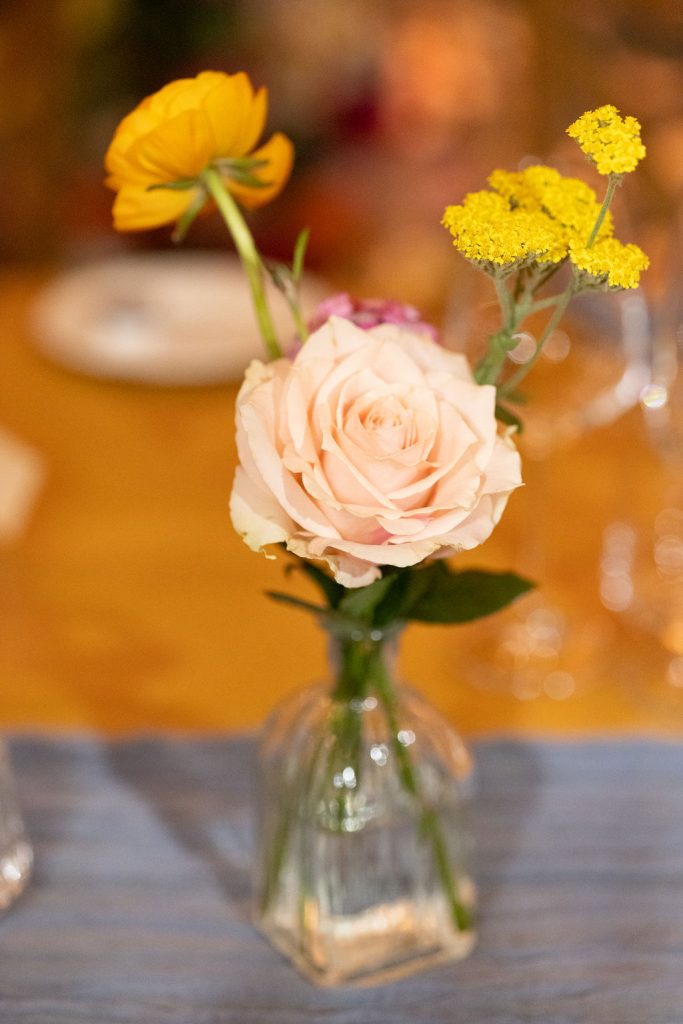 Pink rose in a small clear vase with yellow accent flowers on a striped table.