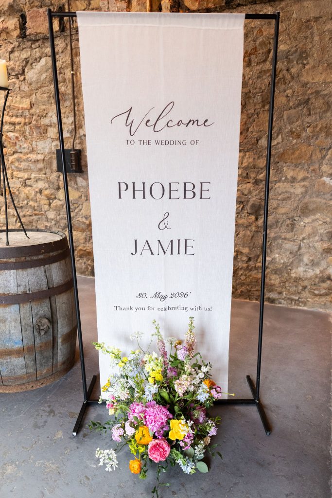 Wedding welcome banner reading 'Phoebe & Jamie', date 30 May 2026, with a colorful floral arrangement at the base.