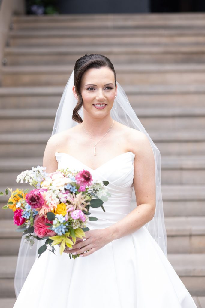Bride in a white strapless wedding gown with a sheer veil, smiling and holding a colorful bouquet of pink, yellow, and blue flowers on stone steps.