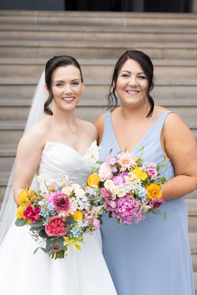 Bride in a white wedding gown with veil stands with a bridesmaid in a pale blue dress, both smiling and holding colorful bouquets on stone steps.