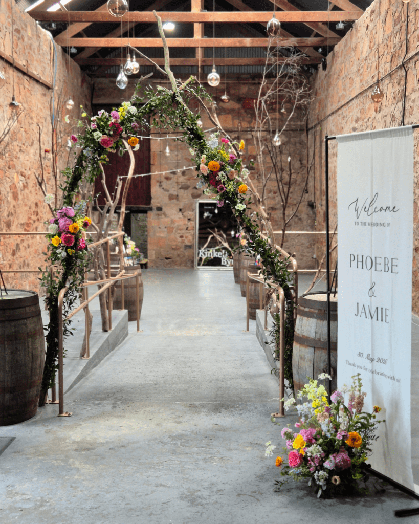 Floral wedding arch in a rustic brick venue, leading to an aisle with barrels and a Welcome banner for Phoebe and Jamie's wedding.