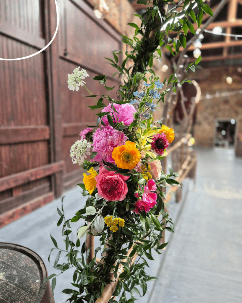 Colorful flower garland wrapped around a wooden railing indoors, with pink peonies, yellow ranunculus, blue blooms, and green leaves.