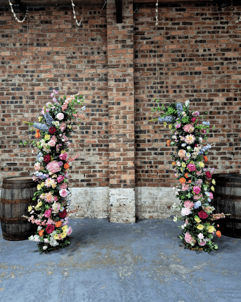 Two tall flower arches with pink, purple, and white blooms against a rustic brick wall, flanked by wooden barrels.