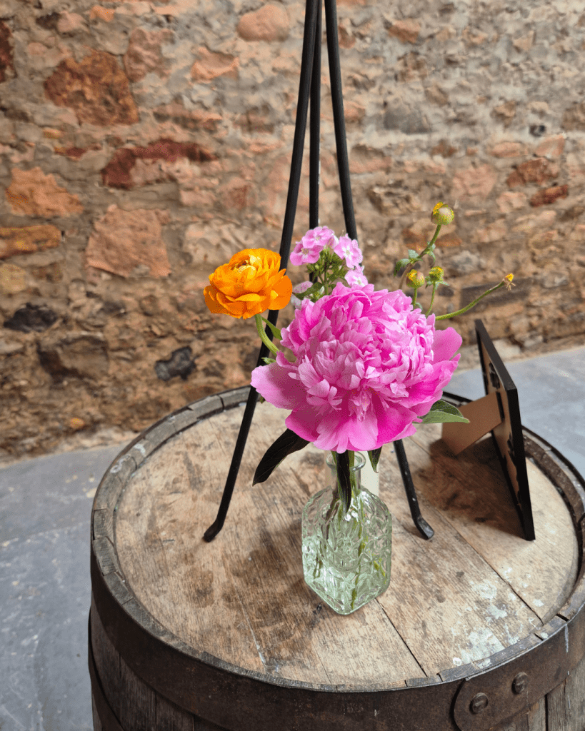 Pink peony and orange ranunculus bouquet in a textured glass vase on a wooden barrel against a brick wall.