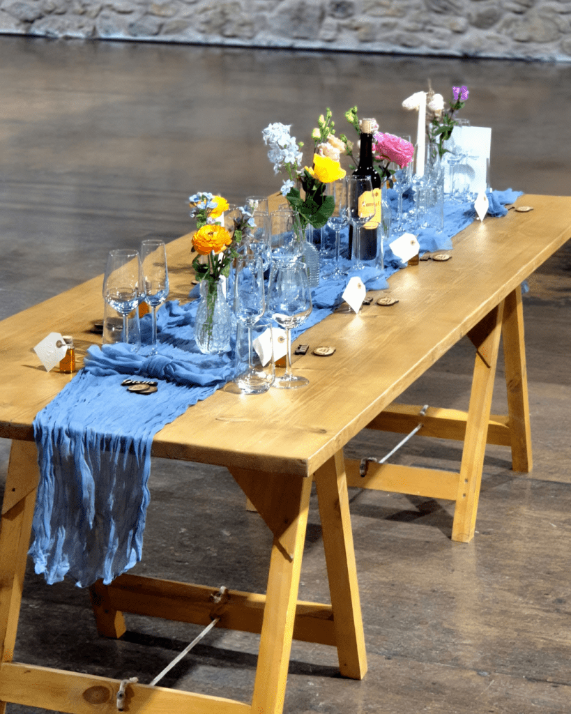 Wooden banquet table with a blue runner, colorful flower vases, wine glasses, and a bottle set for a formal dinner.