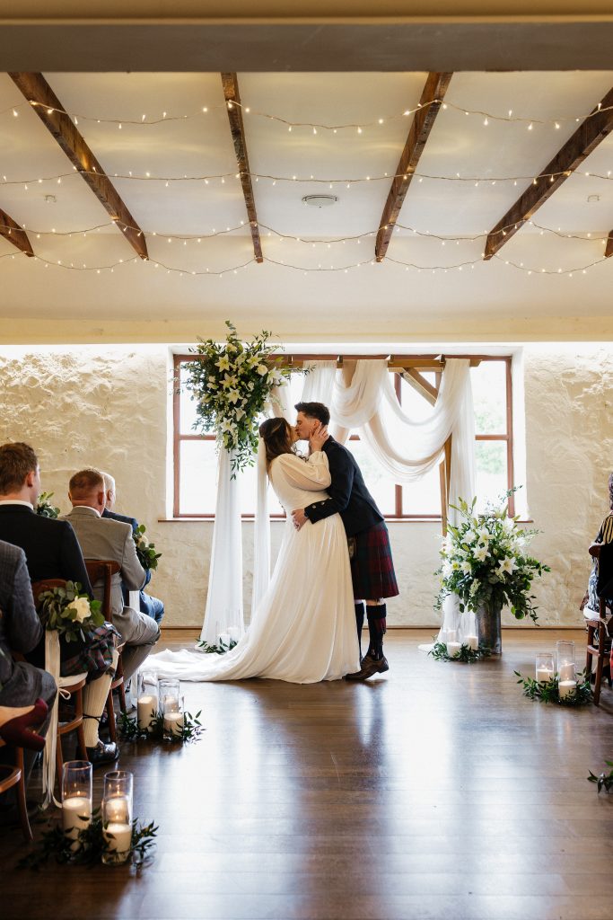 jo paul - fraoch floral design fraoch floral design bride and groom share a kiss at the altar under string lights in a rustic wedding venue with greenery arrangements nearby